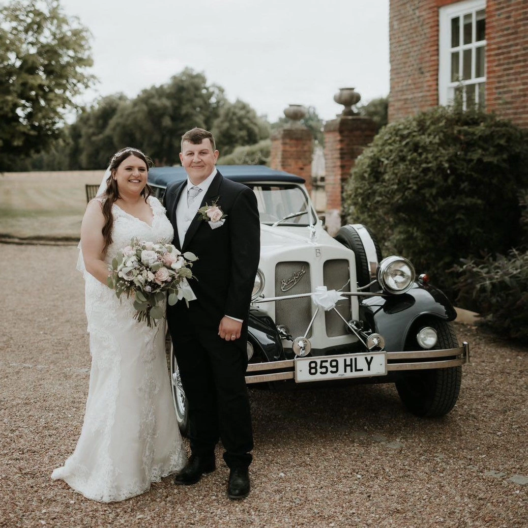 Bride and groom posing in front of a vintage ivory Beauford convertible wedding car at Chilston Park Hotel in Maidstone, Kent, with the bride holding a white floral bouquet beside the classic open top vehicle.