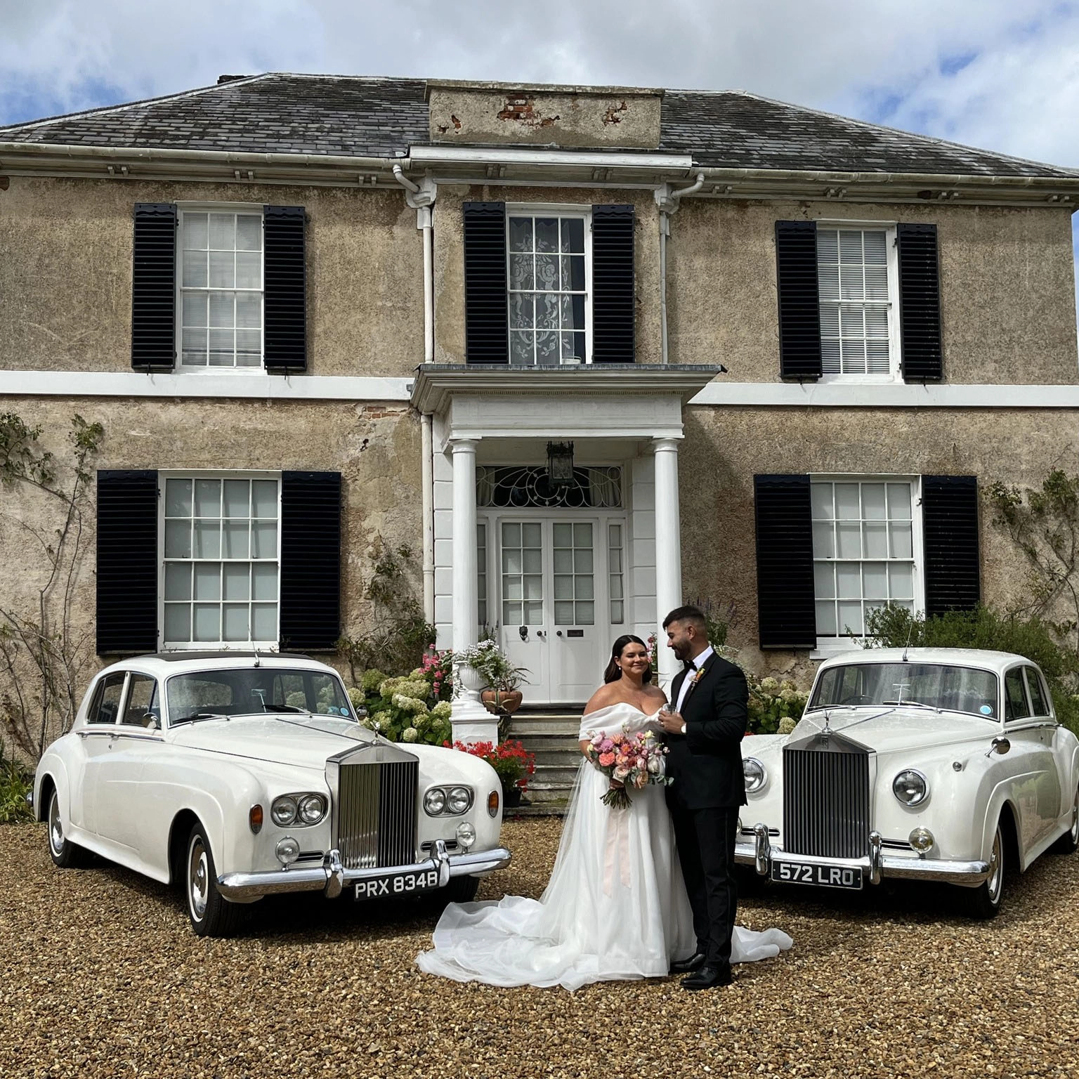Bride and groom standing between two white Rolls-Royce wedding cars outside Preston Court in Canterbury, Kent, with the bride holding a bouquet in front of the elegant country house venue.