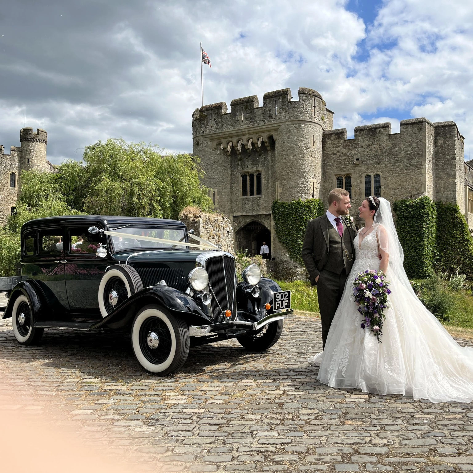 Bride and groom posing beside a black vintage American wedding car at Allington Castle in Medway, Kent, with the historic castle walls and cobbled driveway creating a classic wedding backdrop.