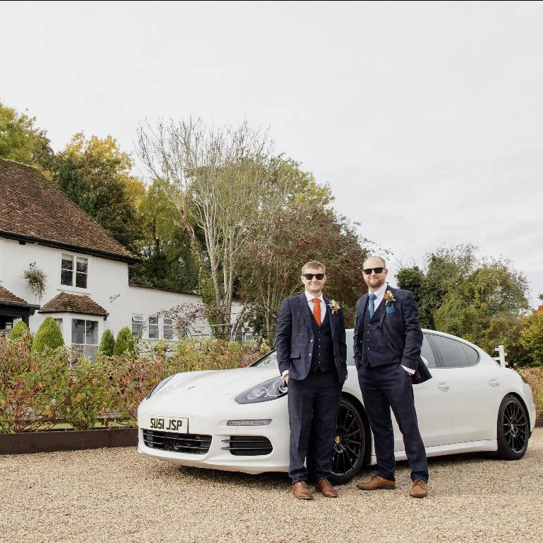 Two groomsmen standing beside a white Porsche wedding car at The Orchard in Otham near Maidstone, Kent, dressed in matching navy suits outside the countryside wedding venue.