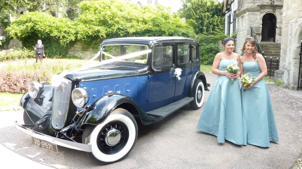 Bridesmaids standing beside blue Austin 18 York vintage wedding car outside church in Kent.