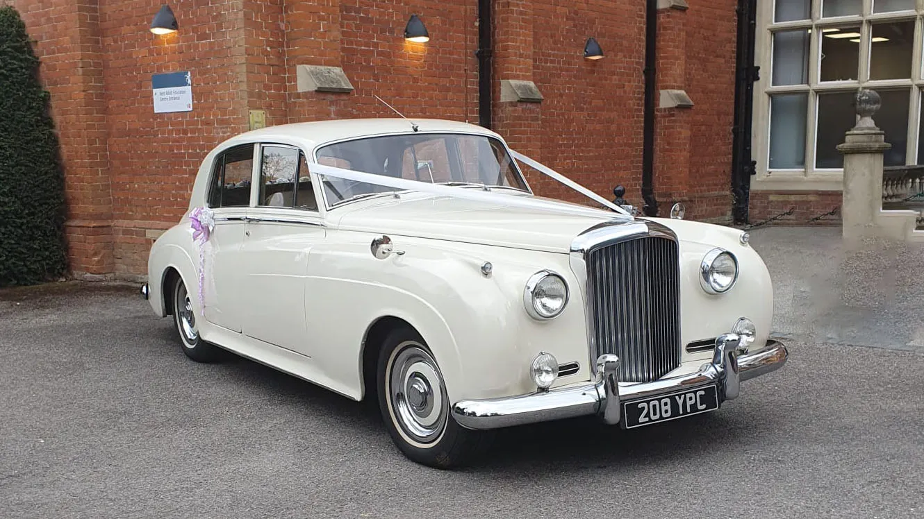 Front three quarter view of a classic Bentley S1 wedding car in Old English White parked outside a church venue in Kent with traditional wedding ribbon decoration.