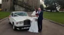 Bride and groom standing beside a classic Rolls Royce Silver Shadow in Old English White outside a historic wedding venue in Kent, with elegant chrome detailing and traditional wedding styling.