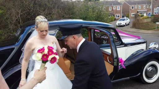Wedding chauffeur helping Bride out of black and Old English White Austin Sheerline wedding car.