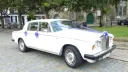 Side view of a white Rolls Royce Silver Shadow wedding car decorated with blue ribbons and floral wedding details, parked outside a church in Kent.