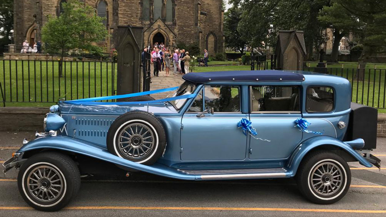Blue vintage Beauford wedding car with Sedanca roof parked outside a church in Kent, featuring cream leather interior and classic wedding styling.