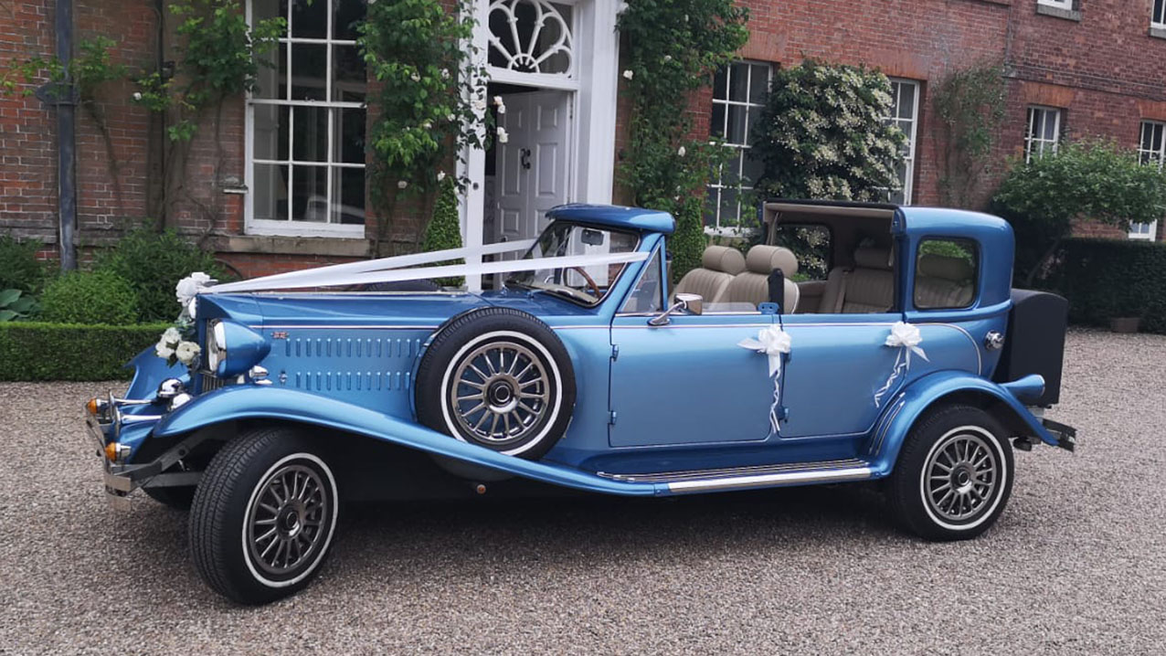 Blue vintage Beauford wedding car with open Sedanca roof outside a wedding venue, decorated with wedding ribbons and chrome detailing.