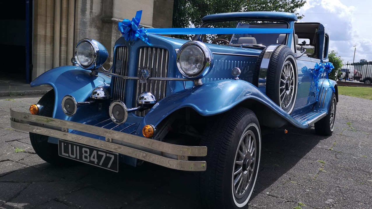 Front view of a blue vintage Beauford wedding car with convertible Sedanca roof, chrome headlights and classic wedding ribbons.