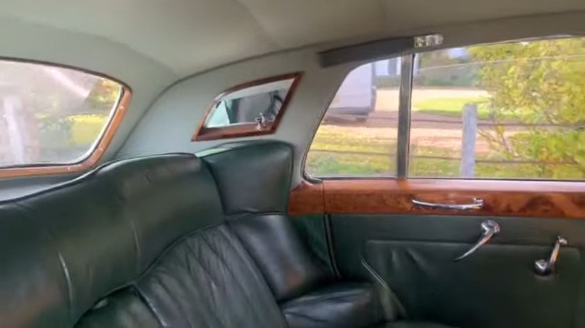 Rear bench seat inside a 1957 Rolls Royce Silver Cloud I finished with dark green leather upholstery and polished wooden picnic tables. Spacious vintage wedding car interior in West Sussex.