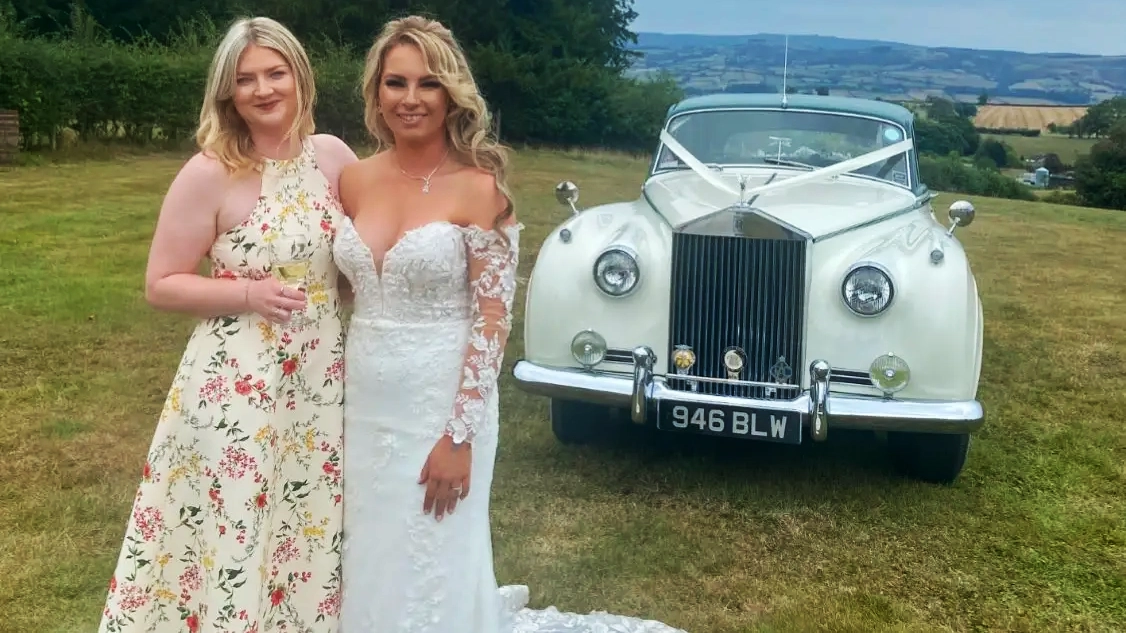 Two brides standing beside a white 1957 Rolls Royce Silver Cloud I with dark green roof at a countryside wedding in West Sussex. Elegant same sex wedding photo with vintage luxury wedding car.