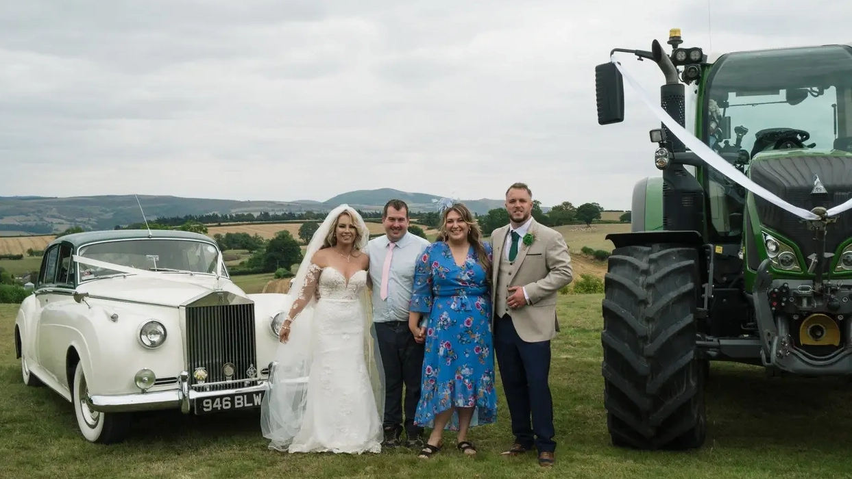 Wedding party posing beside a 1957 Rolls Royce Silver Cloud I and tractor in the West Sussex countryside. White vintage Rolls Royce wedding car with dark green roof at a rural wedding celebration.