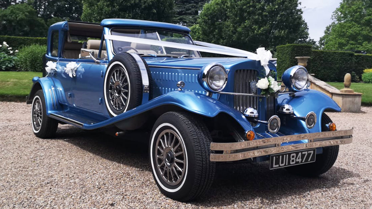 Blue vintage Beauford wedding car decorated with wedding ribbons and bows, photographed outside a countryside wedding venue.