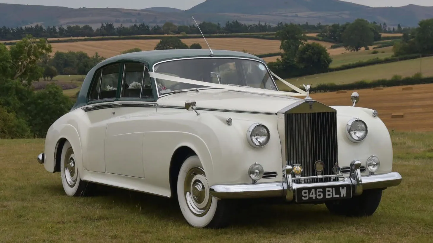 Front three quarter view of a white 1957 Rolls Royce Silver Cloud I with dark green roof in West Sussex. Classic British luxury wedding car with chrome grille and whitewall tyres.