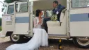Bride and groom sitting inside a classic cream and light blue Volkswagen VW Split Screen Campervan wedding car, with the bride stepping into the rear seating area while the groom smiles from inside. The retro campervan features striped leather seating and floral wedding decorations, photographed outside a church in Lancashire.