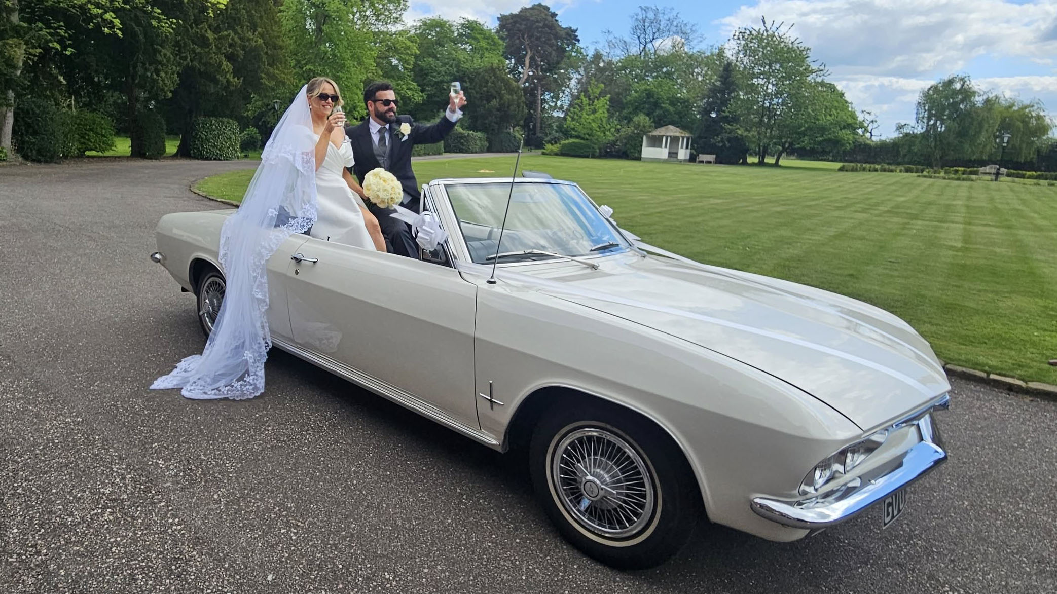 Bride and groom standing beside a classic white Chevrolet Corvair Convertible wedding car with the roof down, photographed in countryside surroundings in Lancashire.
