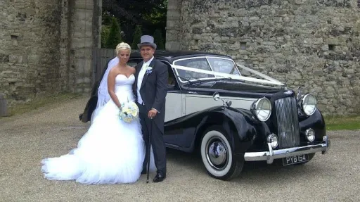 Bride and groom standing beside black and Old English White Austin Sheerline wedding car outside church.
