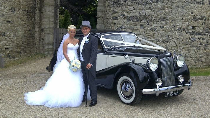 Bride and groom standing beside black and Old English White Austin Sheerline wedding car outside church.