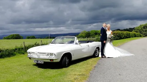 Bride and groom posing beside a white Chevrolet Corvair Convertible wedding car with the roof open under dramatic countryside skies in Lancashire.