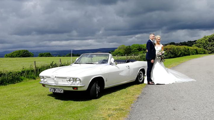 Bride and groom posing beside a white Chevrolet Corvair Convertible wedding car with the roof open under dramatic countryside skies in Lancashire.