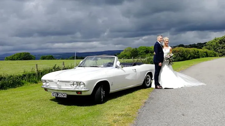 Bride and groom posing beside a white Chevrolet Corvair Convertible wedding car with the roof open under dramatic countryside skies in Lancashire.