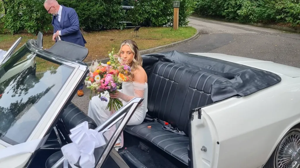 Bride sitting inside a classic white Chevrolet Corvair Convertible with black leather interior and wedding bouquet, while chauffeur assists beside the open top wedding car.