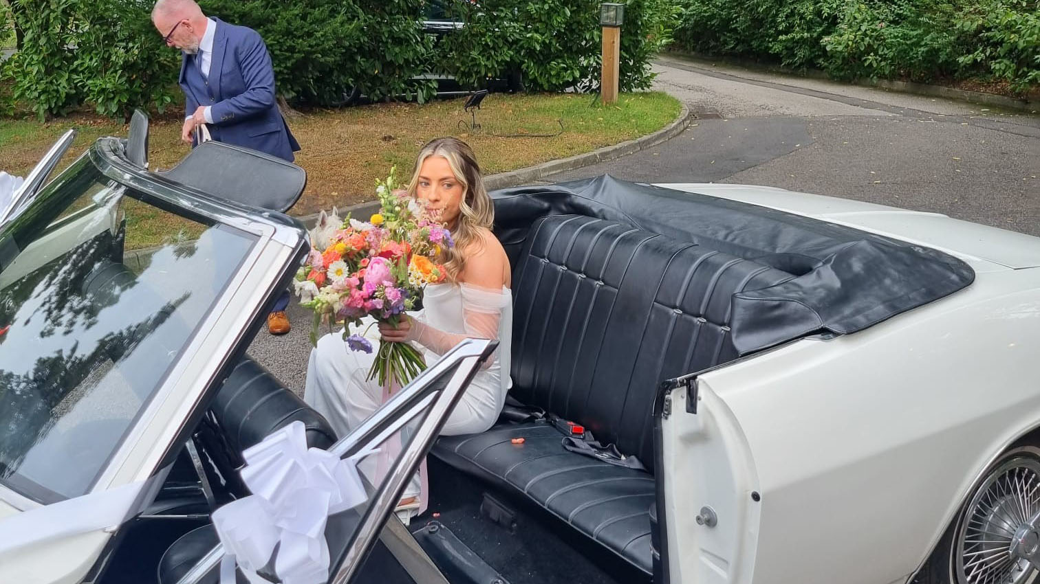 Bride sitting inside a classic white Chevrolet Corvair Convertible with black leather interior and wedding bouquet, while chauffeur assists beside the open top wedding car.