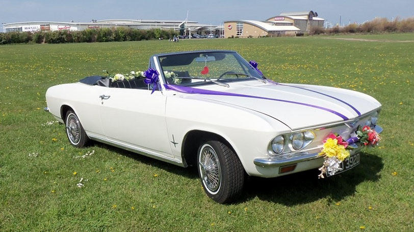 Front three quarter view of a white Chevrolet Corvair Convertible wedding car decorated with purple ribbons and floral wedding arrangements, parked in Lancashire countryside.