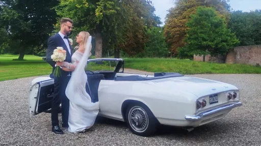 Bride and groom standing beside a classic white Chevrolet Corvair Convertible wedding vehicle with roof folded down, photographed in Lancashire gardens.