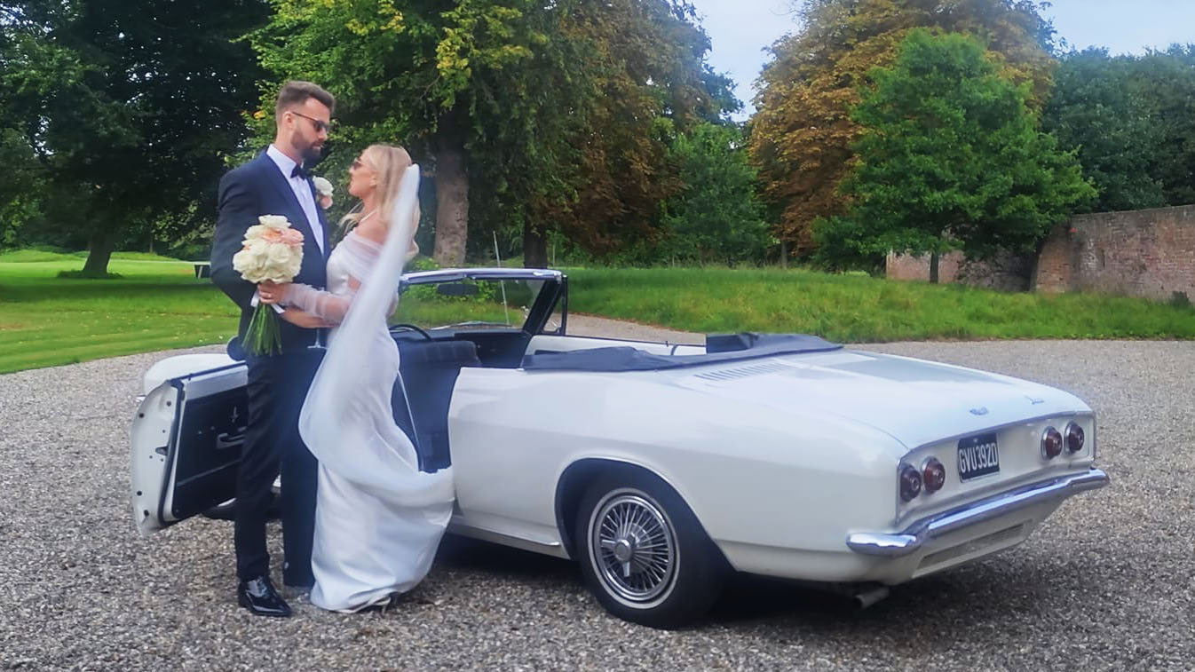 Bride and groom standing beside a classic white Chevrolet Corvair Convertible wedding vehicle with roof folded down, photographed in Lancashire gardens.
