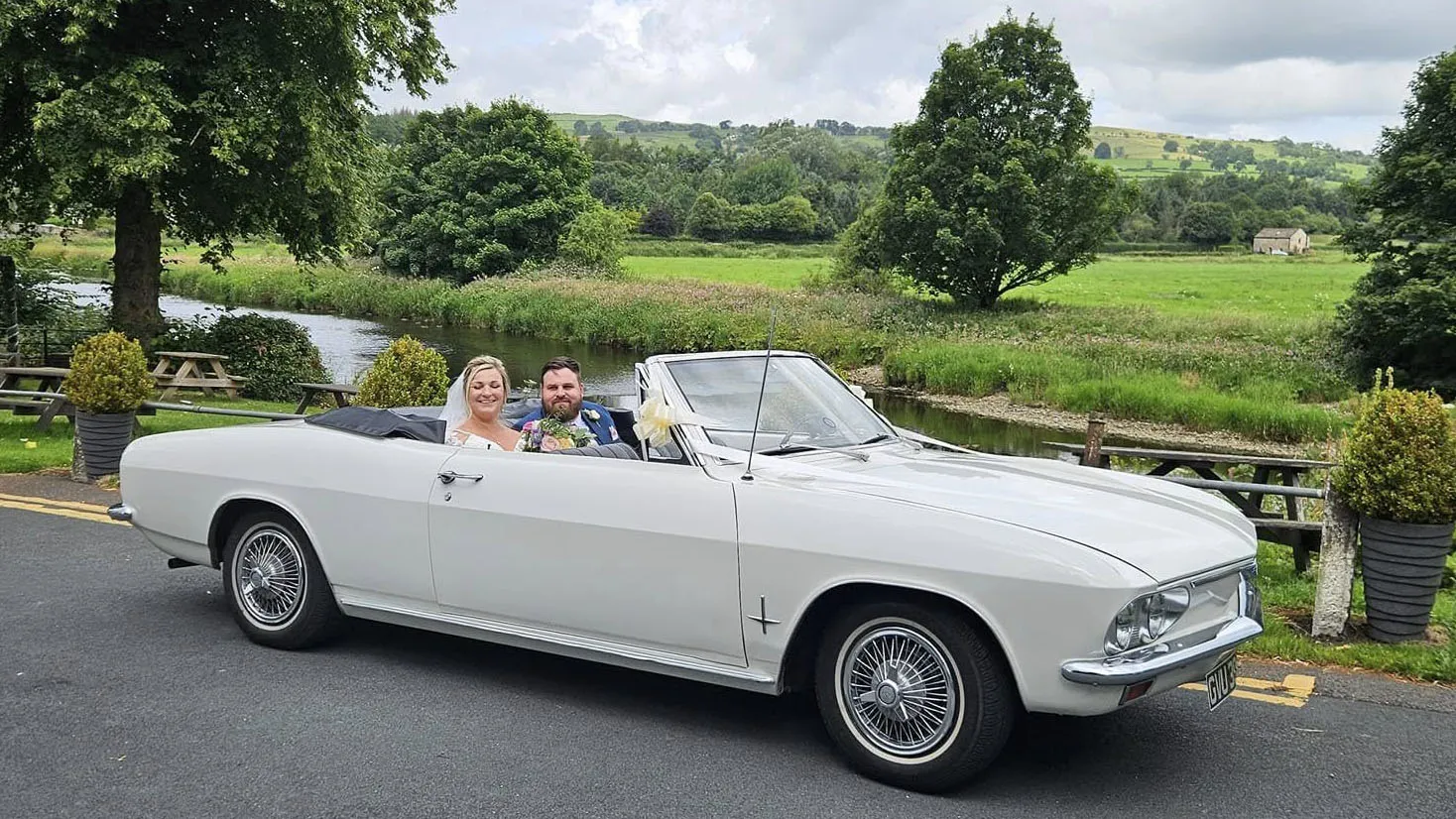 Classic white Chevrolet Corvair Convertible with black roof folded down, carrying wedding passengers through countryside surroundings in Lancashire.