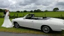 Side profile of a white Chevrolet Corvair Convertible wedding car with open roof, as the bride stands beside the vehicle in a countryside wedding setting.