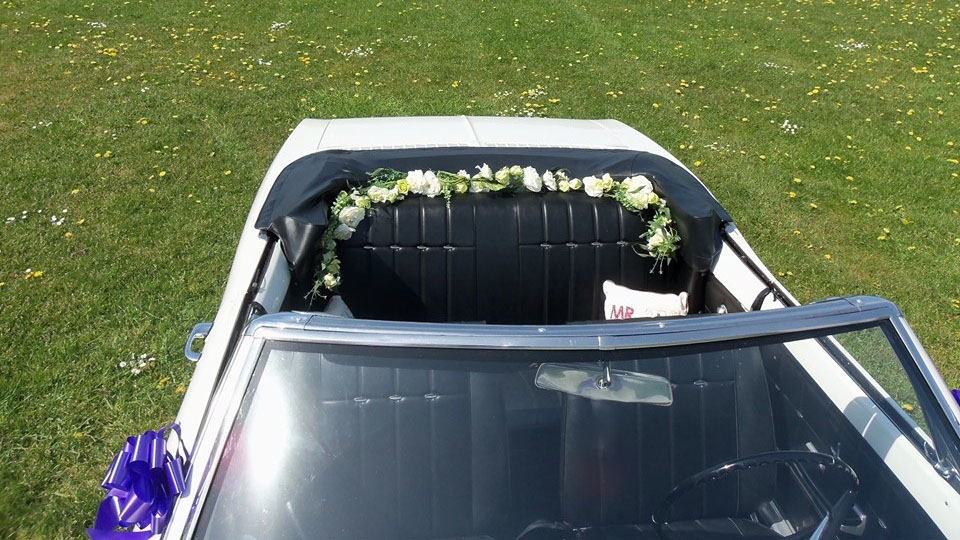 Overhead interior view of a Chevrolet Corvair Convertible with black leather seating, floral garlands and open convertible roof prepared for a wedding ceremony.
