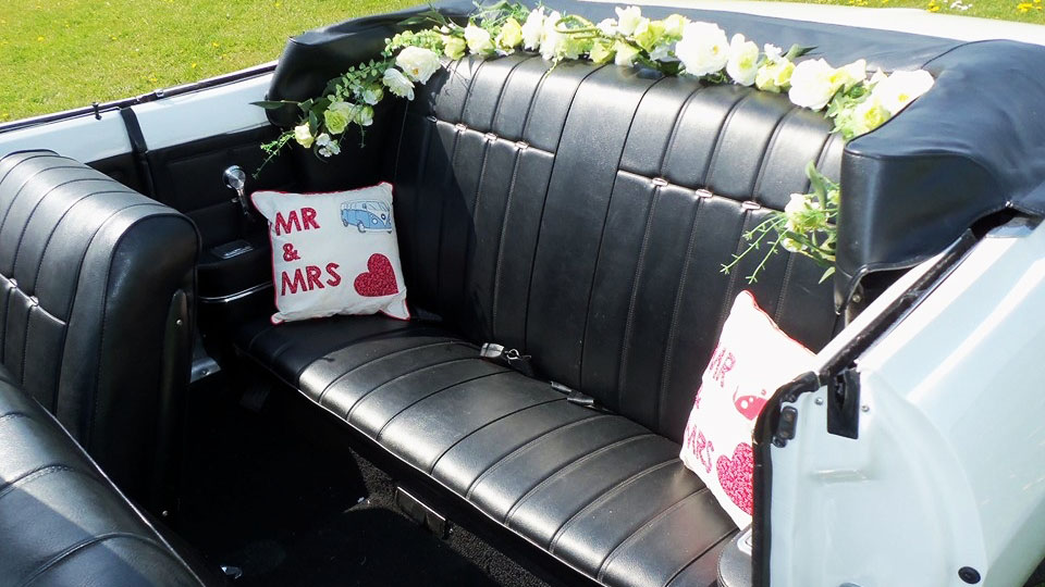 Rear interior seating inside a Chevrolet Corvair Convertible featuring black leather bench seats, wedding floral garlands and personalised Mr & Mrs cushions.