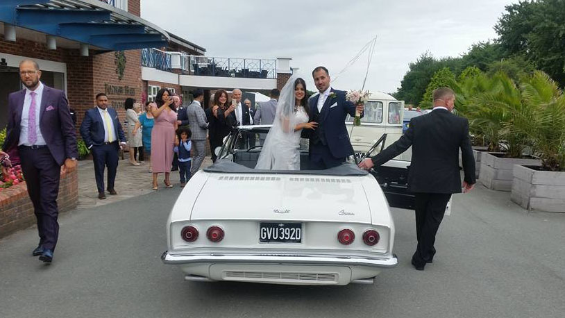 Rear view of a classic white Chevrolet Corvair Convertible wedding car driving away with wedding guests gathered around outside the venue in Lancashire.