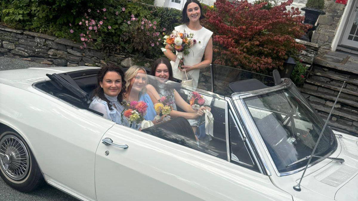 Three bridesmaids seated inside a white Chevrolet Corvair Convertible wedding car with the roof down, smiling and holding bouquets outside a wedding venue.