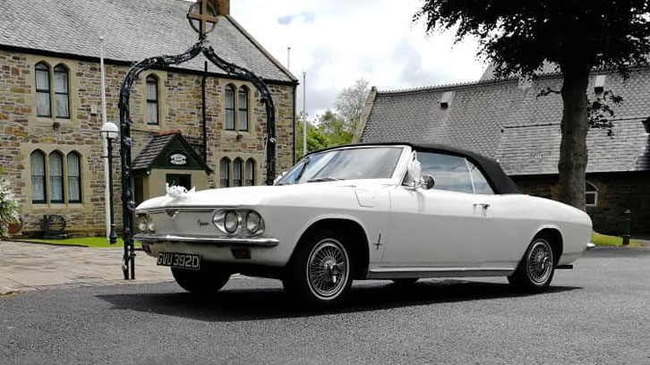 Classic white Chevrolet Corvair Convertible with the black convertible roof raised, parked outside a stone wedding venue in Lancashire.