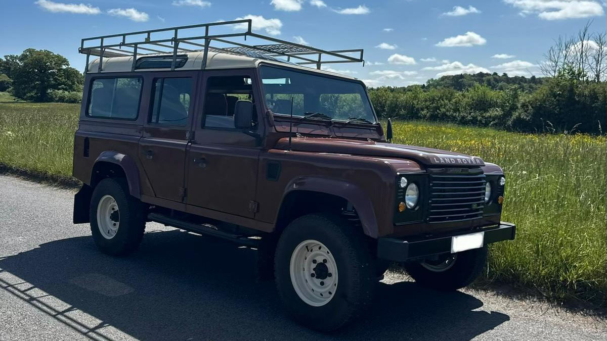 Front side view of brown Land Rover Defender wedding car parked in countryside setting under blue skies.