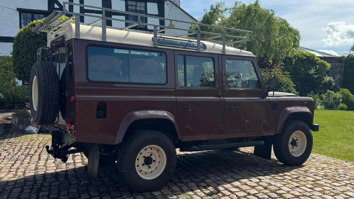 Right side view of brown Land Rover Defender wedding car with roof rack and countryside backdrop.
