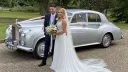 Bride and groom standing beside silver 1960 Rolls-Royce Silver Cloud wedding car at countryside venue.