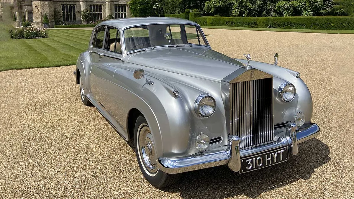 Front side view of silver Rolls-Royce Silver Cloud parked on gravel in Cambridge countryside.