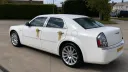 Rear left side view of white Chrysler 300C wedding car with gold ribbons on the door handles, multi-spoke alloy wheels and chrome trim, parked on a driveway with hedges and trees behind.