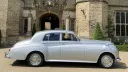 Side profile of silver Rolls-Royce Silver Cloud parked outside historic stone wedding venue.