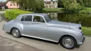 Right side view of silver 1960 Rolls-Royce Silver Cloud wedding car beside countryside lake.