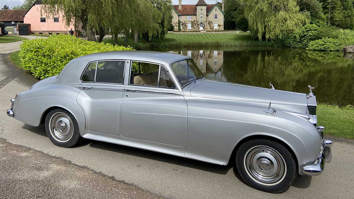 Right side view of silver 1960 Rolls-Royce Silver Cloud wedding car beside countryside lake.