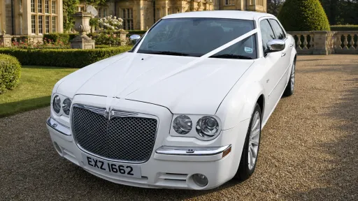 Front view of white Chrysler 300C wedding car, showing chrome grille, headlights and alloy wheels, parked on a gravel driveway.