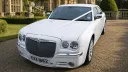 Front view of white Chrysler 300C wedding car, showing chrome grille, headlights and alloy wheels, parked on a gravel driveway.