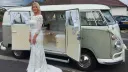 Bride standing beside ivory and sage green Volkswagen Split Screen campervan with side doors open in Devon.