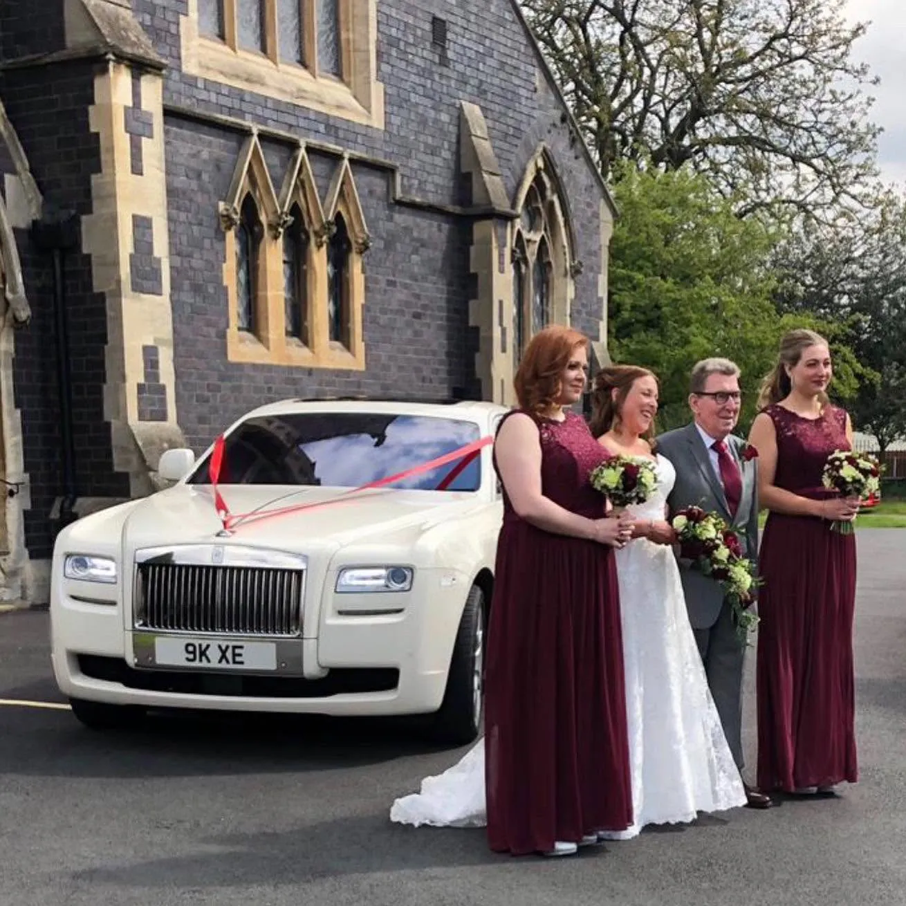 White Rolls-Royce Ghost wedding car with bride in her white wedding gown and her bridesmaids wearing burgundy dresses outside a church in the West Midlands.