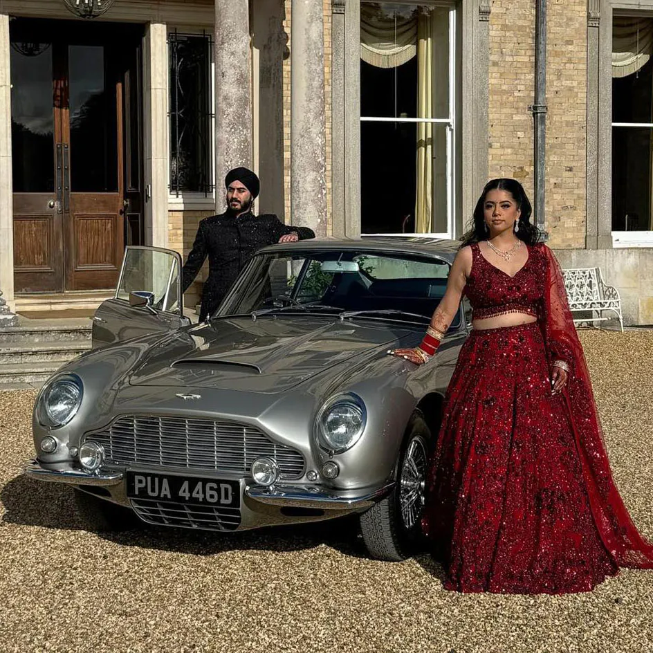Asian bride and groom posing with a classic Aston Martin wedding car outside a grand wedding venue.
