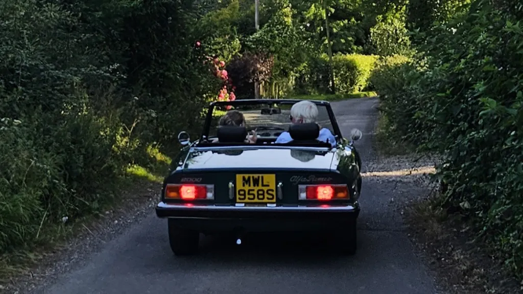 Rear view of green Alfa Romeo Spider Veloce 2000 convertible driving along country lane with roof down.